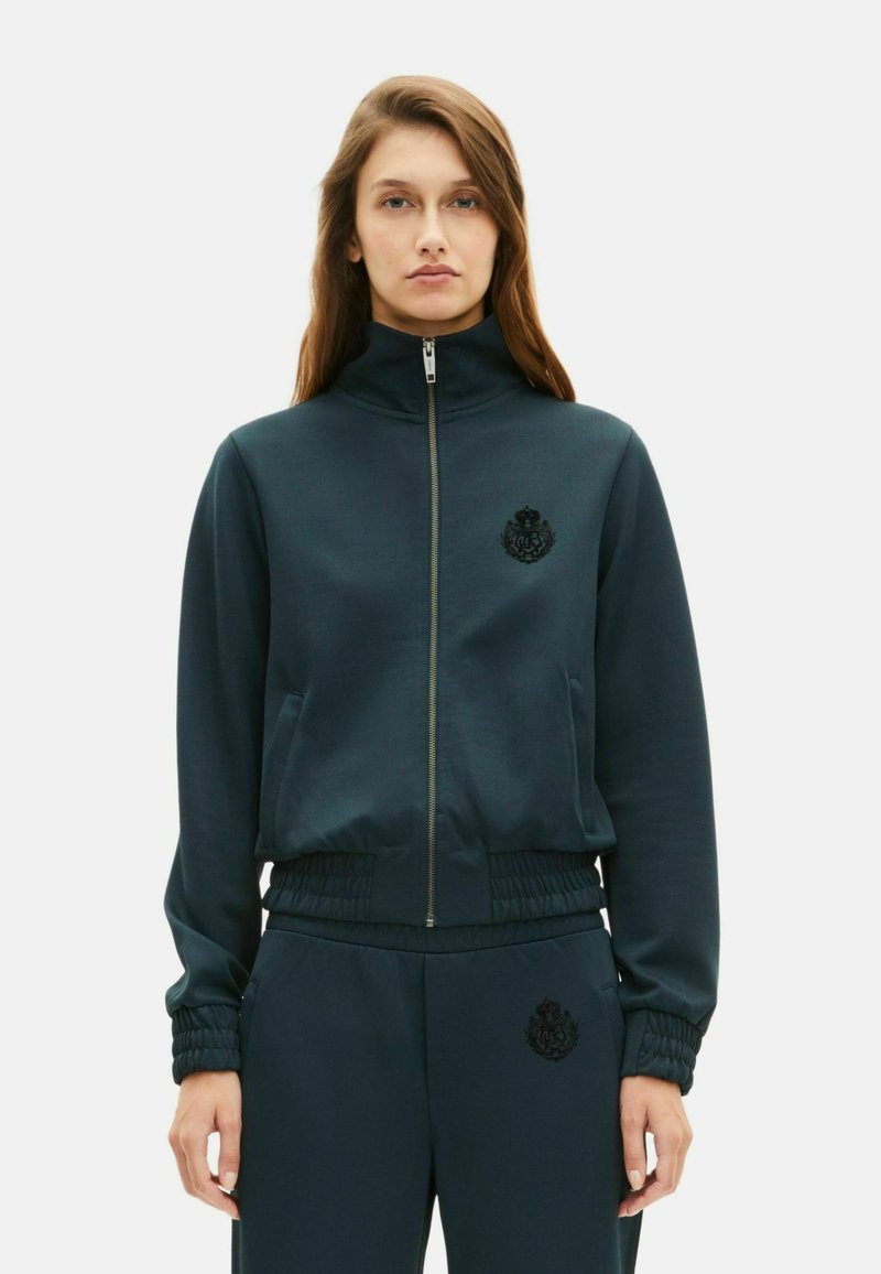 Marineblauwe ritsjack met hoge kraag, elastische manchetten en tailleband. Bevat een zwart geborduurd logo op de borst. Glad textuur van de stof.