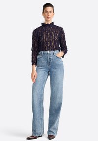 Una modella posa con le mani in tasca, indossando un top in pizzo trasparente blu navy e jeans a vita alta di colore azzurro chiaro, abbinati a stivali con punta affusolata.