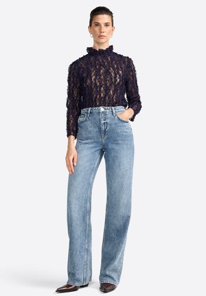 A model poses with hands in pockets, wearing a sheer navy lace top and light blue high-waisted jeans, complemented by pointed-toe boots.