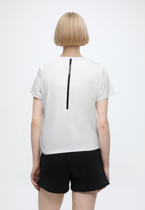 Person med kort blond hår vendt væk, iført en hvid T-shirt med en sort lodret stribe og sorte shorts mod en ensfarvet baggrund.