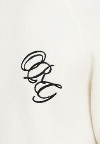 Weißer Pullover mit weichem Material, der ein schwarzes gesticktes Monogramm "ORG" in einem eleganten Schreibschrift-Design zeigt.
