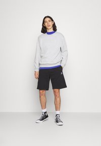 Grauer Sweatshirt mit gerippten Bündchen, über einem blauen T-Shirt getragen; schwarze Shorts mit Logo; schwarze Sneakers mit weißen Akzenten. Leichter, lässiger Stil.