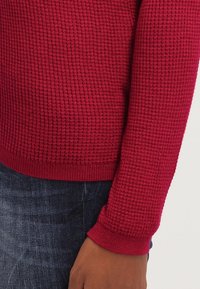 Rode gebreide trui met een gestructureerd patroon, geribbelde mouwen en zoom, gecombineerd met donkere spijkerbroek. Het materiaal lijkt zacht en warm.