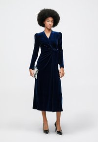 Ženska z afrom nosi dolgo temno modro žametno obleko z zavojem, črne pete in drži srebrno torbico, stoji pred enobarvno podlago.