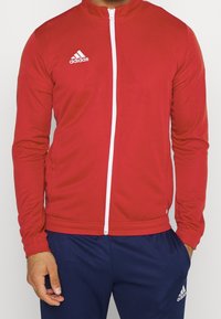 Veste de sport zippée rouge en tissu lisse et extensible. Présente une fermeture éclair blanche et un logo Adidas sur la poitrine gauche.