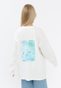 Witte lange mouwen shirt met een groot lichtblauw abstract waterontwerp op de achterkant. Zachte stof en ontspannen pasvorm.