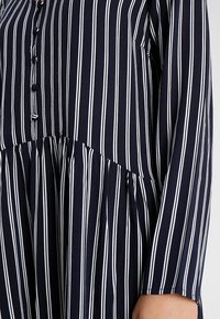 Granatowa sukienka z guzikami, w pionowe białe paski, z długimi rękawami i marszczoną talią. Miękka tkanina o lekkiej strukturze.