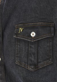Dunkelblaue Jeanshemd mit einer Brusttasche. Mit gelber "JV" Stickerei, silbernem Knopf und kontrastierendem orangefarbigem Nähten. Weiche Textur.
