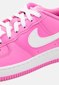 Vaaleanpunaiset synteettisestä nahasta valmistetut tennarit valkoisilla yksityiskohdilla, joissa on pyöreä kärki, valkoiset nauhat ja teksturoitu pohja. Nike-logo sivussa.