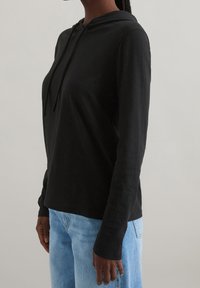 Persona che indossa una felpa nera a maniche lunghe abbinata a jeans di denim blu chiaro, in piedi di lato contro uno sfondo neutro.