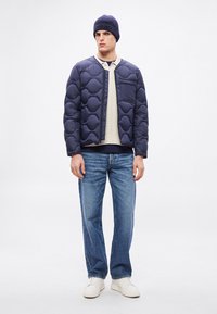 Μπλε σκούρο καπιτονέ μπουφάν με φερμουάρ μπροστά, οριζόντιο σχέδιο, τσέπη στο στήθος, φορεμένο πάνω από ελ αφρύ πλεκτό πουλόβερ και μπλε τζιν, ολοκληρωμένο με λευκά αθλητικά παπούτσια.