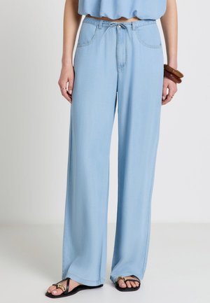 Pantalon large bleu clair avec poches avant et bouton, porté avec des sandales noires et un top bleu court.