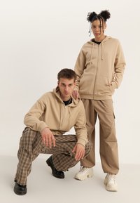 Felpe beige con zip e cappuccio, abbinate a pantaloni beige e a quadri. Il modello indossa sneaker bianche e nere. Lo sfondo è bianco uniforme.