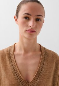 Suéter marrón de cuello en V con un borde acanalado, de textura ligera, llevado por una persona. El fondo es completamente blanco.