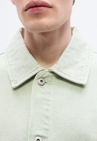 Ανοιχτό πράσινο τζιν μπουφά ν με εντυπωσιακό μεγάλο γιακά, δύο ασημένια κουμπιά και διακριτικό υφή στο ύφασμα. Έμφαση στις λεπτομέρειες του γιακά και των κουμπιών.