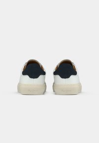 Baskets blanches avec accent talon bleu marine foncé, corps en tissu texturé, semelle en caoutchouc et détails subtils de logo à l'arrière. Design classique à tige basse.