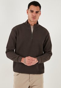 Pullover zip-up marrone a coste con colletto alto, dotato di maniche lunghe e polsini elasticizzati. Il tessuto ha una texture morbida e una vestibilità rilassata.