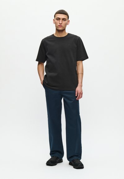 Tricou negru cu mânecă scurtă, croială lejeră, asociat cu pantaloni largi de culoare bleumarin. Încălțăminte neagră cu suprafață texturată. Fundal alb.