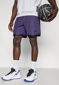 Violet basketbola šorti ar melnu apdari, teksturēts sieta materiāls. Baltas sporta apavi ar ziliem akcentiem, kopā ar baltām garajām zeķēm.