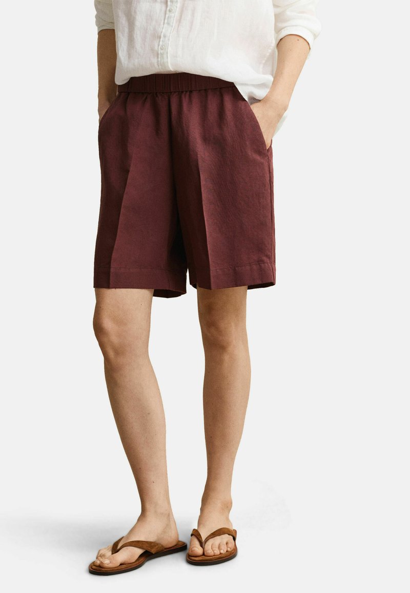 Persoon draagt bordeauxrode knielange shorts, wit shirt met lange mouwen opgerold, en bruine teenslippers, handen in zakken.
