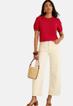 Top rosso a maniche corte con bordi in pizzo, abbinato a pantaloni a vita alta cream a gamba larga. Tiene una borsa di paglia beige e indossa sandali con tacco marroni.