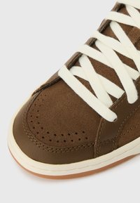 Sapatilha de couro castanho e camurça com atacadores brancos, bico redondo, perfurações na caixa dos dedos e sola de borracha texturizada.