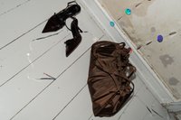 Barna szatén táska összegyűrt tetejjel, párosítva fekete hegyes orrú magassarkú cipőkkel. A háttérben színes díszítő elemek láthatók egy fa padlón.