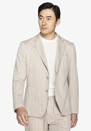 Mężczyzna w beżowej marynarce w prążki i dopasowanych spodniach, w białej koszulce z okrągłym dekoltem, patrzący nieco w prawo.