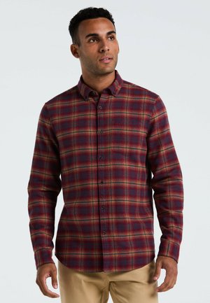 Man in een rood en blauw geruit overhemd met knopen en beige broek, staand met ontspannen handen, iets omhoog en opzij kijkend.