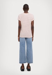 Osoba v světle růžovém tričku s krátkým rukávem, širokých modrých džínách s roztrženými lemy, černých platformových botách a zlatých kruhových náušnicích, otočená zády.