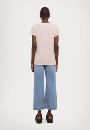 Persona che indossa una camicia rosa chiaro a maniche corte, jeans a gamba larga blu con orli sfilacciati, scarpe nere con plateau e orecchini a cerchio dorati, di spalle.
