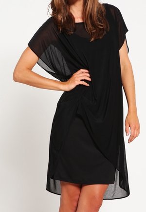 Kobieta w czarnej sukience do kolan z przezroczystymi, luźnymi krótkimi rękawami oraz asymetryczną nakładką, stoi z jedną ręką na biodrze.