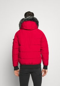 Rød pufferjakke med hætte i grå pels, med en quiltet design og ribbede manchetter. Sorte jeans fuldender outfittet.
