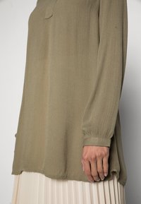 Haut vert olive à manches longues en tissu texturé, coupe droite avec une poche avant discrète, associé à une jupe plissée crème.