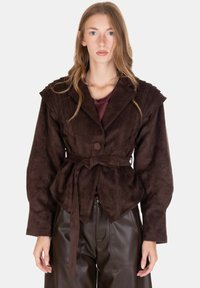 Giacca in suede marrone con cintura a fiocco, spalle strutturate, maniche larghe e dettagli testurizzati sulla parte anteriore. Indossata sopra una maglietta color granato.