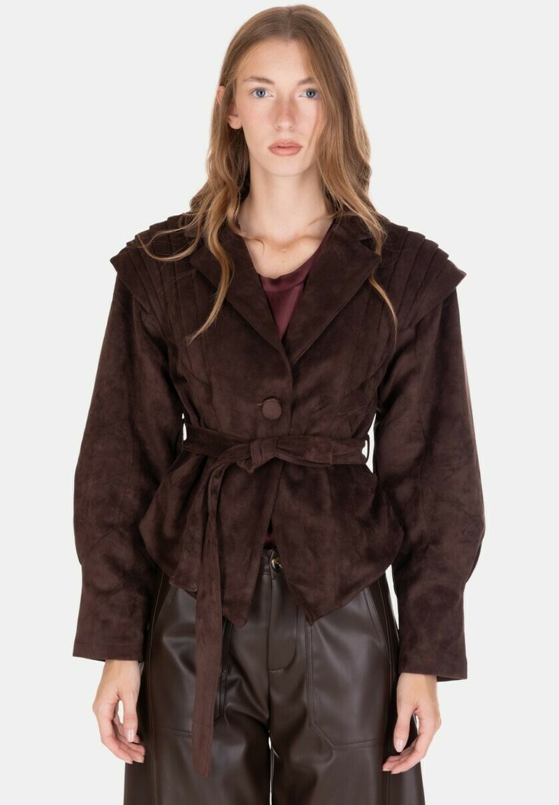 Giacca in suede marrone con cintura a fiocco, spalle strutturate, maniche larghe e dettagli testurizzati sulla parte anteriore. Indossata sopra una maglietta color granato.