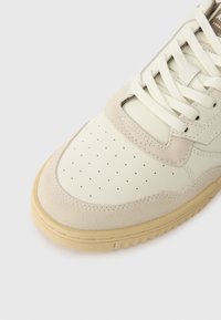 Basket beiger avec boîte à orteils perforée, accents en daim, lacets crème et semelle en caoutchouc marron clair sur fond blanc.