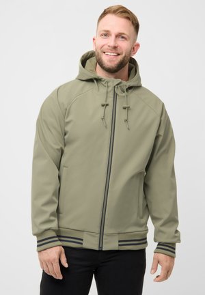 Lächelnder Mann, der eine hellolivfarbene, mit Reißverschluss versehene Kapuzenjacke mit gestreiften Bündchen und schwarze Hosen trägt, vor einem schlichten weißen Hintergrund stehend.