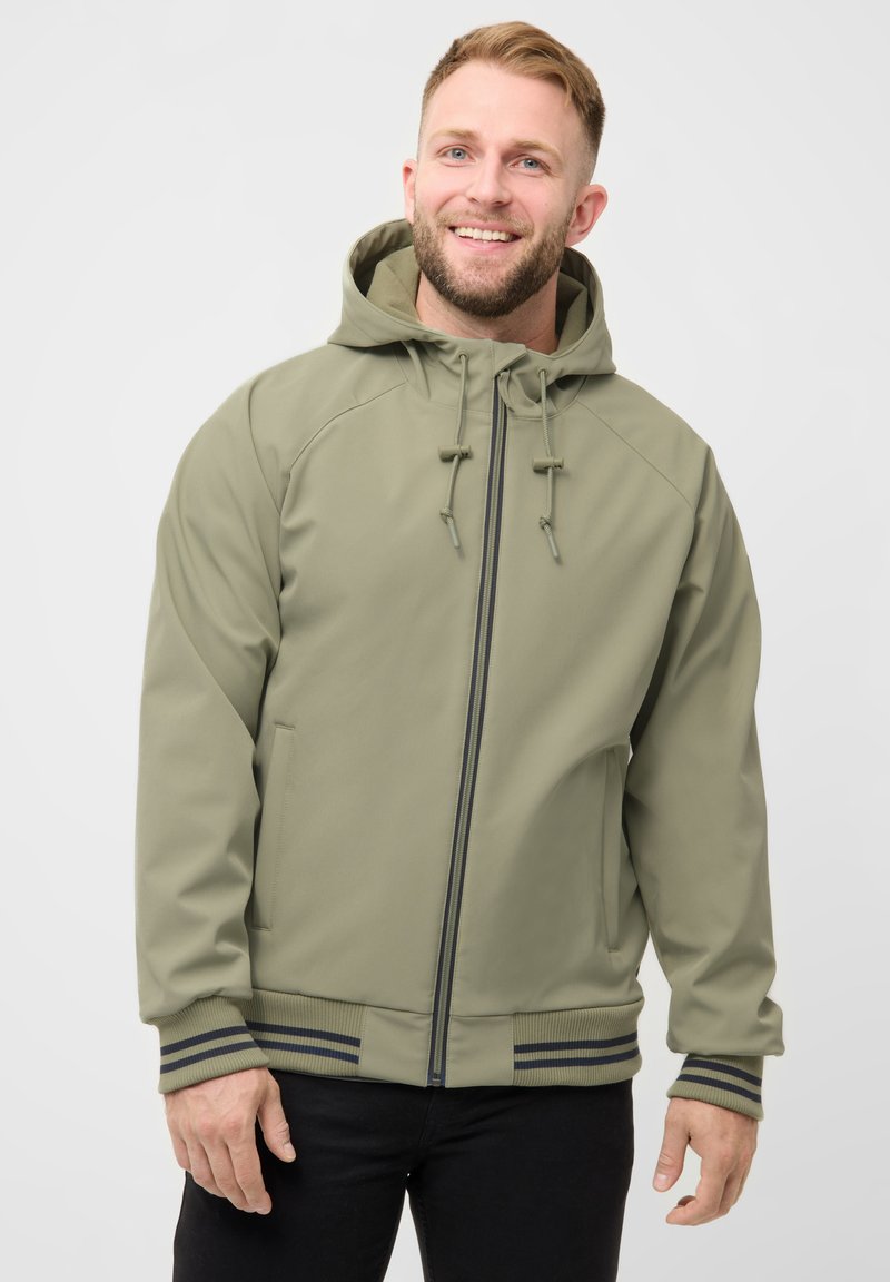 Lächelnder Mann, der eine hellolivfarbene, mit Reißverschluss versehene Kapuzenjacke mit gestreiften Bündchen und schwarze Hosen trägt, vor einem schlichten weißen Hintergrund stehend.