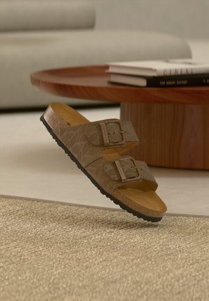 Brune mønstrede sandaler med to justerbare spænder, korkfodseng og struktureret sort gummisål, vist over en beige vævet tæppe.