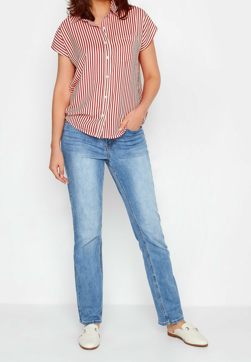 Chemise à manches courtes à rayures rouges et blanches avec des boutons, associée à un jean bleu clair et des chaussures blanches faciles à enfiler, présentant une coupe décontractée et un détail de poche.