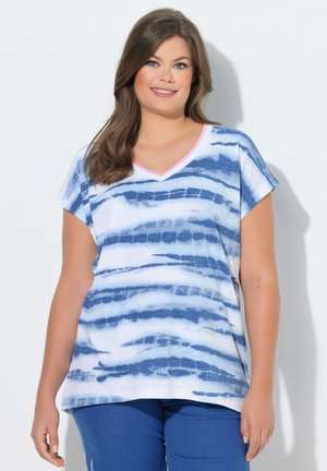 Uśmiechnięta kobieta o długich brązowych włosach nosi białą i niebieską koszulkę w paski z dekoltem w serek oraz niebieskie spodnie, stojąc na białym tle.