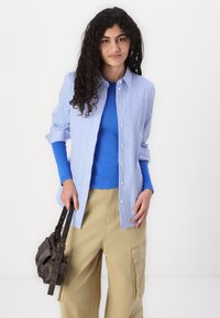 Chemise à rayures bleues et blanches avec un haut bleu à manches longues en dessous, associée à un pantalon cargo taille large beige et un sac à bandoulière marron.