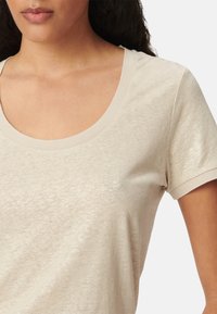 Vrouw in een beige t-shirt met korte mouwen en een ronde hals, weergegeven van schouders tot midden torso tegen een effen achtergrond.