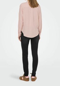 Camisa de manga larga rosa claro con dobladillo curvo y pliegue en la parte trasera, combinada con jeans negros y zapatillas marrones con detalles en rosa.