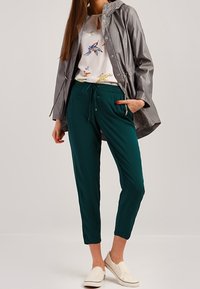 Giacca impermeabile grigia leggera, top bianco con fiori, pantaloni verdi a sigaretta e scarpe beige slip-on. Presenta vita con cordoncino e cappuccio.