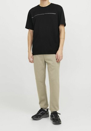 Personne portant un t-shirt noir avec une fine bande argentée et un pantalon beige, debout en baskets noires sur un fond blanc uni.