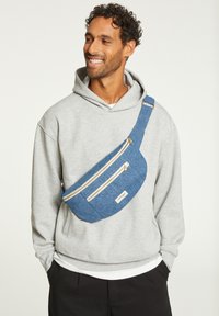 Sac banane en tissu bleu avec trois poches zippées et une étiquette blanche. Porté en bandoulière avec un sweat à capuche gris et un pantalon noir.