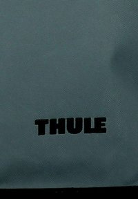 Logo de la marque Thule imprimé en lettres noires en gras sur un fond de tissu texturé gris.