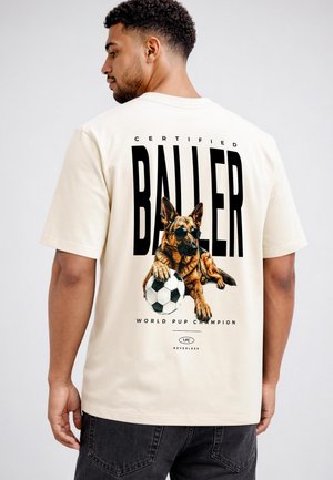 Man wearing a beige t-shirt with a graphic of a German Shepherd dog in sunglasses holding a soccer ball and the words "Certified Baller."